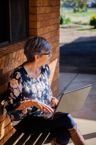 Mature woman sitting on park bench chair outside working on laptop in sunshine - Australian Stock Image