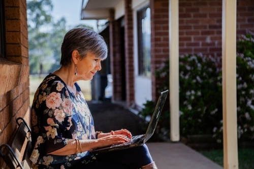 Mature woman sitting on park bench chair outside working on laptop in sunshine - Australian Stock Image