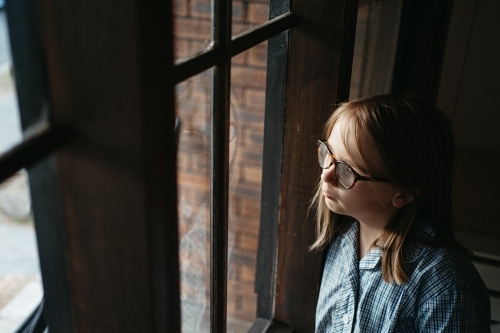 Melancholic primary aged girl in a school uniform looks out a window - Australian Stock Image