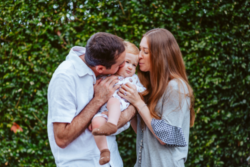 mid aged mother and father squishes young baby boy with blond hair  in front of a green background - Australian Stock Image