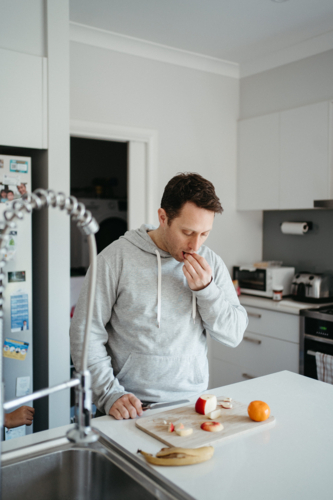 Middle-aged guy eating and cutting fruit on the kitchen counter. - Australian Stock Image