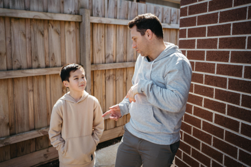 Middle-aged man making funny face with a boy whos hands are in his hoodie pocket. - Australian Stock Image