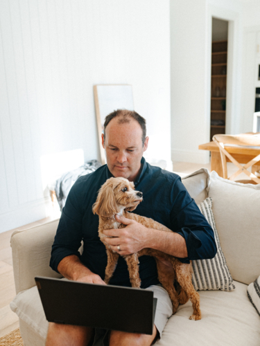 Middle aged man working on his laptop and holding a small dog while sitting on the couch. - Australian Stock Image