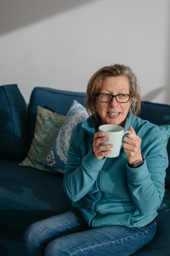 Middle aged woman sitting on couch, drinking hot beverage - Australian Stock Image