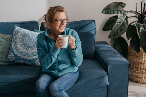 Middle aged woman sitting on couch, drinking hot beverage - Australian Stock Image
