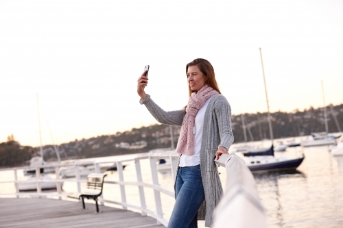 Middle aged woman using mobile phone on wharf to take selfie - Australian Stock Image
