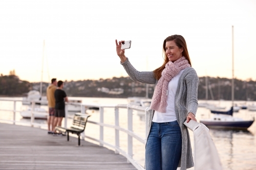 Middle aged woman using mobile phone on wharf to take selfie - Australian Stock Image