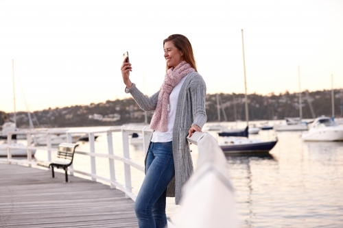 Middle aged woman using mobile phone on wharf to take selfie - Australian Stock Image