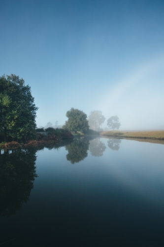Misty morning beside a river with calm water, reeds and trees - Australian Stock Image