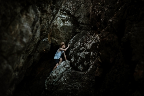 Moody image of young boy wearing hat climbing in dark cave in tropical north Queensland - Australian Stock Image