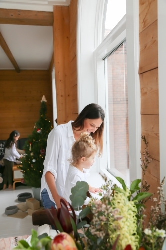 Mother and daughter looking out the window with girl decorating Christmas tree in background - Australian Stock Image
