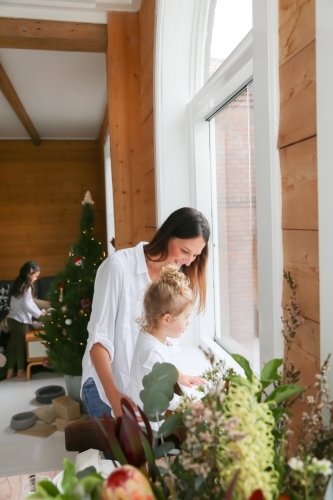 Mother and daughter looking out the window with girl decorating Christmas tree in background