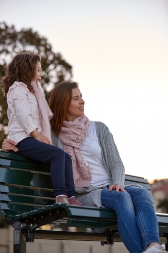 Mother and daughter sitting on park bench at park on sunset - Australian Stock Image