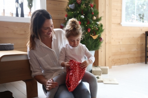 Mother and daughter unwrapping present with Christmas tree in background - Australian Stock Image