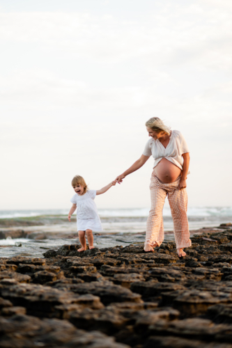 Mother and daughter walking along rocky coastline at sunset, expectant mother smiling joyfully - Australian Stock Image