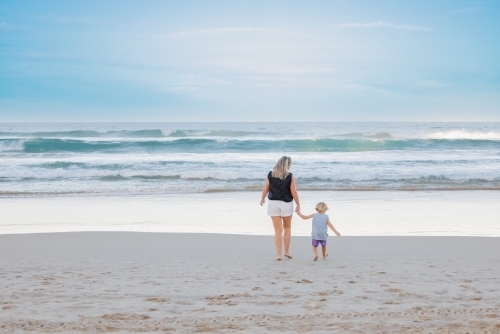 Mother and son holding hands walking towards water on the beach - Australian Stock Image