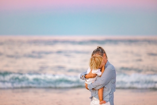 Mother and toddler son embracing on the beach with pink sunset sky - Australian Stock Image