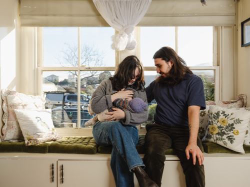 Mother breastfeeding baby wearing beanie on window seat with father sitting beside them - Australian Stock Image