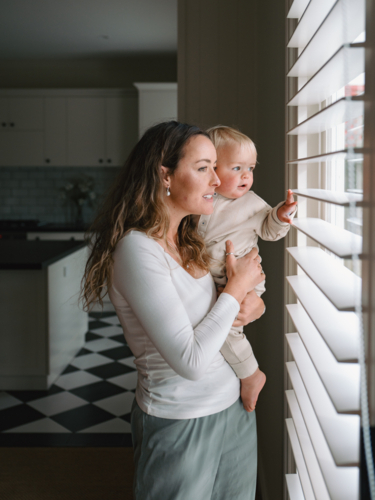 Mother carrying baby peeking through window blinds - Australian Stock Image