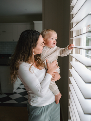 Mother carrying baby peeking through window blinds - Australian Stock Image