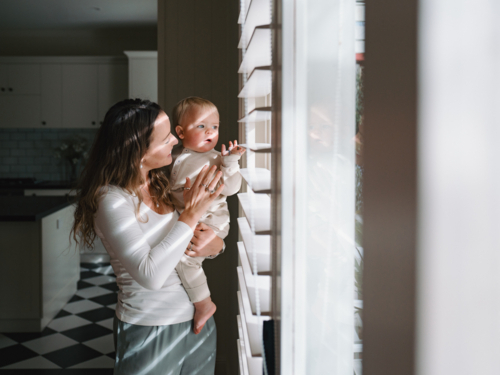 Mother carrying baby peeking through window blinds - Australian Stock Image