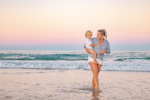 Mother carrying son on the beach in pink twilight light. Sunset on the Gold Coast. - Australian Stock Image