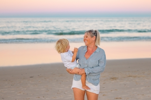 Mother carrying son on the beach in pink twilight light. Sunset on the Gold Coast. - Australian Stock Image