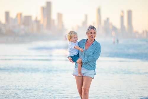Mother carrying son on the beach with Gold Coast city skyline in background - Australian Stock Image