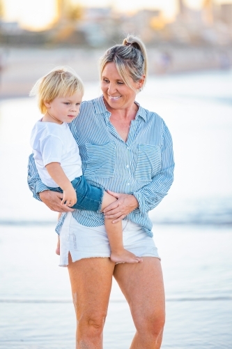 Mother carrying son on the beach with Gold Coast city skyline in background - Australian Stock Image