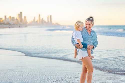 Mother carrying son on the beach with Gold Coast city skyline in background - Australian Stock Image