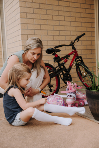 Mother crouching beside her daughter putting on her socks. - Australian Stock Image