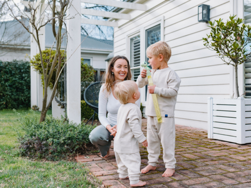 Mother crouching on brick patio playing bubbles with two sons - Australian Stock Image