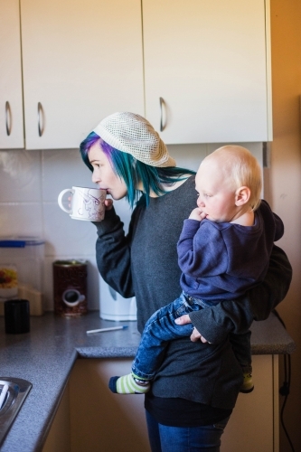 Mother drinking from cup while holding child on hip - Australian Stock Image