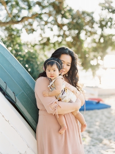 Mother holding baby girl at the beach, leaning against some boats - Australian Stock Image