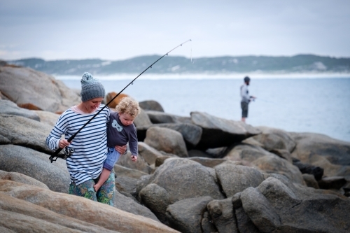 Mother holding daughter walking through rocks on beach with fisherman in background coastal backdrop - Australian Stock Image