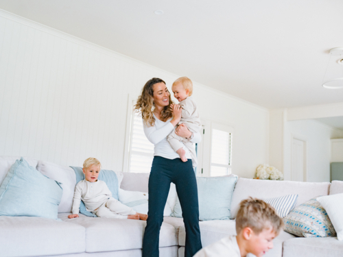 Mother holding toddler baby with young boys playing around her in the living room - Australian Stock Image
