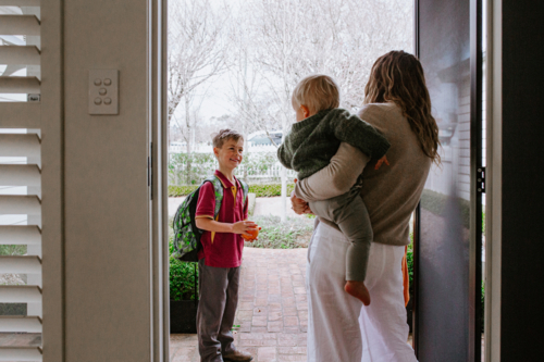 Mother holding toddler son in her hip saying goodbye to older son with backpack out the doorway - Australian Stock Image