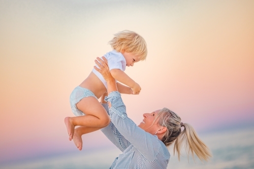 Mother lifting toddler son in the air on the beach with pink sunset sky - Australian Stock Image