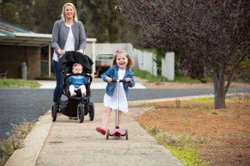 Mother pushing baby in pram and young daughter on scooter - Australian Stock Image