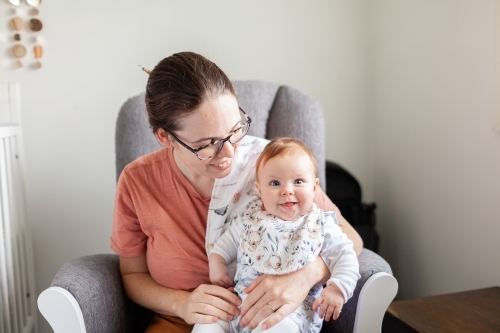 Mother sitting in rocking chair in nursery with delighted baby smiling and cuddling together - Australian Stock Image