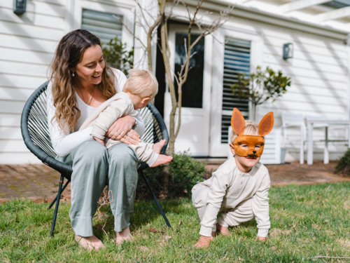 Mother sitting on black woven chair cradling baby boy with child on ground wearing kangaroo mask - Australian Stock Image