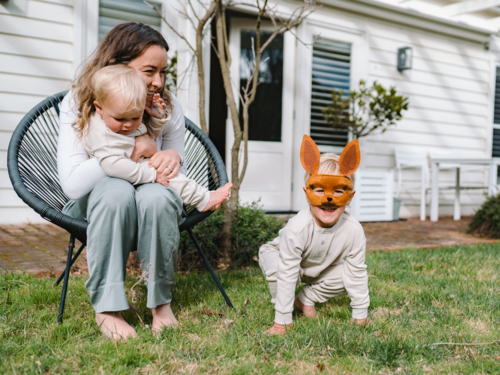 Mother sitting on black woven chair cradling baby boy with child on ground wearing kangaroo mask - Australian Stock Image