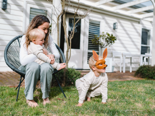 Mother sitting on black woven chair cradling baby boy with child on ground wearing kangaroo mask - Australian Stock Image
