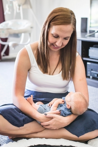 Mother sitting on floor at home smiling down at newborn baby in arms - Australian Stock Image