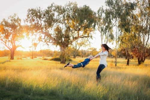 Mother spinning son in field with long grass at sunset - Australian Stock Image