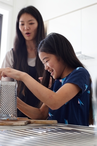 Mother watching over daughter whilst using the grater in kitchen - Australian Stock Image