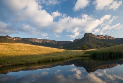 Mountain escarpment, sunlit grass and clouds reflected in farm dam
