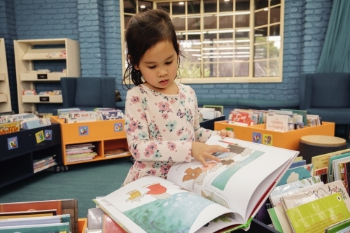 Multicultural child reading a book in library - Australian Stock Image