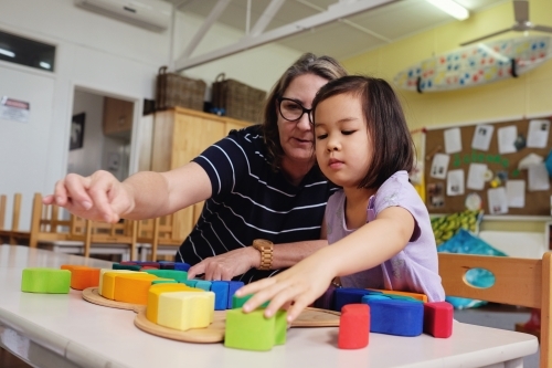 Multicultural teacher and child playing wooden blocks puzzles in kindergarten - Australian Stock Image
