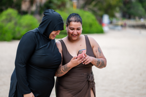 Multiracial women checking their phone on the sandy shore at the beach - Australian Stock Image